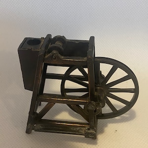 May include: A vintage metal pencil sharpener shaped like a small wooden wagon with a wheel. The sharpener is made of dark metal and has a small, rectangular opening for pencils. The wheel is made of metal and has spokes.