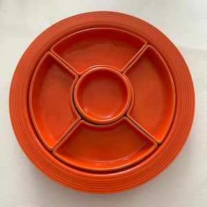 May include: A bright orange ceramic serving platter featuring a large circular base with concentric ring detailing. The platter includes four removable curved wedge-shaped dishes and one central circular bowl, all in a matching vibrant orange glaze.