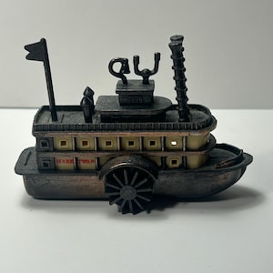 May include: A miniature metal paddle steamer boat model featuring a dark bronze finish, a central paddle wheel, and a cream-colored cabin section. The side of the vessel is clearly labeled with the text MARK TWAIN in small red lettering.