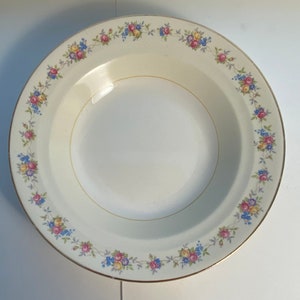 May include: A white ceramic soup bowl with a gold rim and a floral pattern around the edge. The flowers are pink, yellow, and blue.