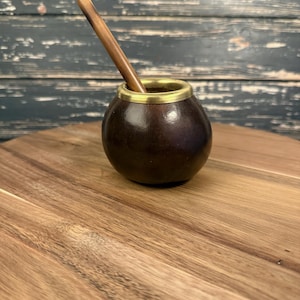 Könnte beinhalten: Eine dunkelbraune, kürbisförmige Yerba-Mate-Tasse mit einem goldfarbenen Rand und einem Holzstrohhalm. Die Tasse steht auf einer Holzoberfläche. Der Strohhalm ist in die Tasse gesteckt. Der Hintergrund ist dunkles Holz.