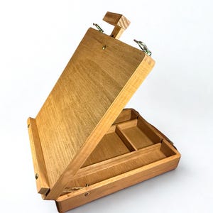 Könnte beinhalten: Eine hölzerne Künstlerbox mit einem Klappdeckel und eingebauten Fächern. Die Box ist aus hellfarbenem Holz gefertigt und hat eine natürliche Oberfläche. Der Deckel wird mit einer Holzstütze offen gehalten. Die Box ist für die Aufbewahrung von Kunstbedarf konzipiert.