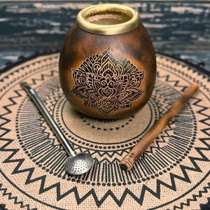 May include: A brown gourd with a gold rim and a black mandala design. The gourd is sitting on a brown and black woven mat with a straw bombilla and a metal bombilla.