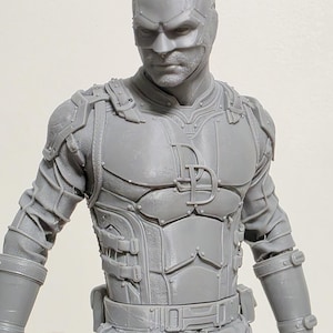 May include: A gray action figure of a superhero in a detailed costume. The figure has a mask with horns, a chest emblem, and armor-like detailing. The figure is in a standing pose, showcasing the intricate design of the outfit.
