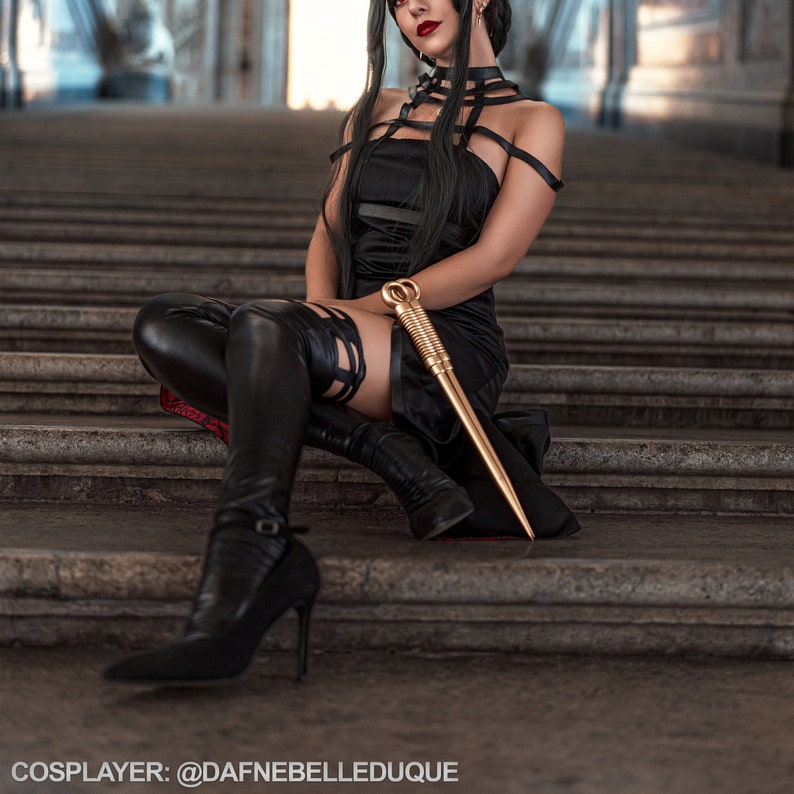 Peut inclure: Une femme en robe noire et bottes cuissardes pose avec un b&acirc;ton dor&eacute;. Elle a les cheveux noirs et le rouge &agrave; l&egrave;vres rouge. L'arri&egrave;re-plan est un ensemble de marches en pierre.