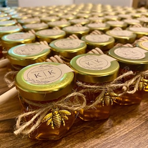 May include: A collection of small glass jars filled with honey, each with a gold lid and a personalized label with the initials "K.K." and the date "09.29.2024". Each jar is tied with twine and decorated with a small gold bee charm.