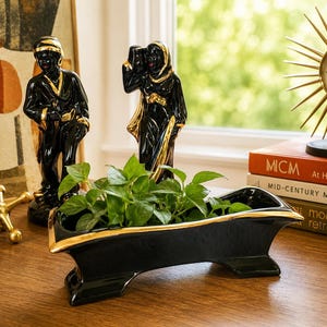May include: A black ceramic planter with a gold-trimmed rim and base, featuring a long, rectangular, footed design. The planter is filled with a vibrant green leafy plant, showcasing a glossy finish that reflects light along its edges.
