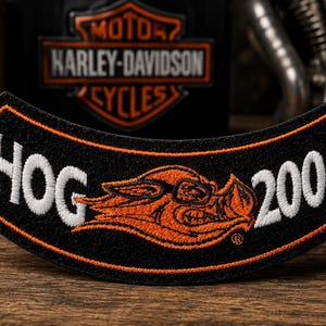 May include: A curved, black embroidered patch featuring the white text HOG on the left and 2006 on the right. In the center, an orange and black embroidered graphic of a stylized hog head wearing sunglasses is displayed with an orange border.