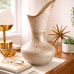 May include: A decorative white vase with gold accents. The vase has a unique shape with a wide opening and a textured surface. The gold trim adds a touch of elegance. The vase is a stylish home decor item.