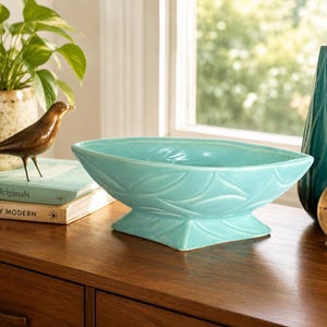 May include: A turquoise ceramic bowl with a pointed, elongated oval shape and a square pedestal base. The exterior surface of the bowl features a repeating embossed leaf pattern that wraps around the sides and the base of the vessel.