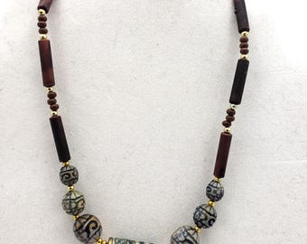 May include: A handcrafted necklace featuring a mix of dark brown, cylindrical wooden beads, small gold-colored beads, and patterned, spherical beads in shades of green and gray. The necklace has a central, patterned cylindrical bead.