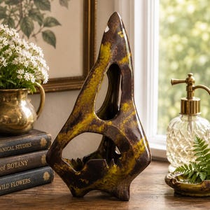 May include: A tall, abstract ceramic sculpture featuring a pointed, triangular silhouette with two large, irregular cutouts. The surface is finished in a mottled glaze of deep chocolate brown and vibrant chartreuse yellow, creating a textured appearance.