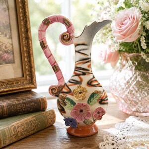 May include: A ceramic pitcher featuring a white body with black and orange abstract swirling lines, a pink handle with gold accents, and three-dimensional floral embellishments in pink, blue, and gold. The base is a solid orange color.