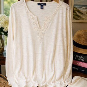 May include: A long-sleeve cream-colored top with a split V-neckline featuring intricate white lace trim. The shirt has a soft, textured fabric and a Chaps brand label at the inner collar. The garment is displayed on a wooden hanger.