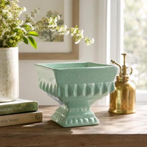 May include: A square, mint green ceramic pedestal bowl featuring a textured, fluted design along the upper rim and a stepped base. The bowl has a glossy, crackled glaze finish and sits centered, showcasing its symmetrical, footed structure.
