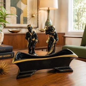 May include: A black ceramic planter with a long, boat-like shape and gold-painted trim along the top edges. The planter features a wide, open center and stands on two flared feet, showcasing a glossy finish that reflects light across its surface.