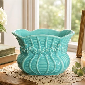May include: A turquoise ceramic planter with a scalloped rim and a basket-weave texture. The exterior features a horizontal band of embossed rose motifs encircling the middle section, with vertical ribs extending from the base to the rim.