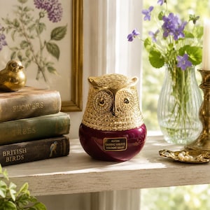 May include: A decorative owl-shaped container with a metallic gold textured head and a deep burgundy base. A rectangular label on the front reads AVON RAINING VIOLETS COLOGNE GELEE. The object is a vintage-style vanity item for fragrance storage.