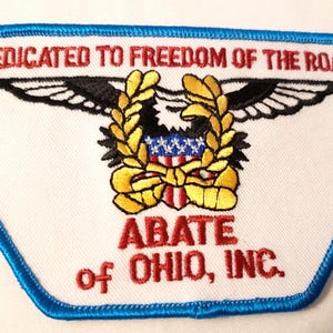 May include: A white embroidered patch with a blue border featuring a black eagle with spread wings, a gold wreath, and a shield with red, white, and blue stripes. The text reads DEDICATED TO FREEDOM OF THE ROAD and ABATE OF OHIO, INC. in red.