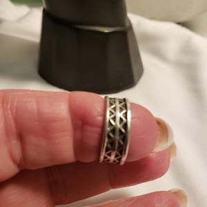 May include: A silver ring with a geometric pattern. The ring features a series of triangles and other shapes. The ring is shown on a finger.