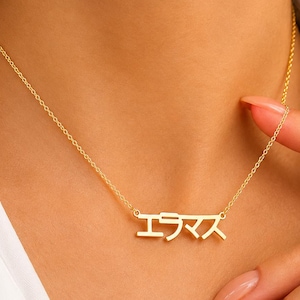 May include: A gold-toned necklace featuring a delicate chain and a central pendant displaying the Japanese katakana characters エラマス. The metal has a polished finish, and the pendant is suspended directly from the chain links, creating a simple design.