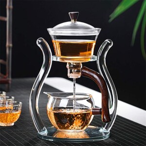 May include: A clear glass tea infuser with a wooden handle and accents. The infuser has a top chamber with a lid, a middle chamber, and a bottom chamber. Tea is being poured from the top chamber into the bottom chamber. The tea is amber in color.