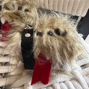 May include: Two fluffy, furry, gray and white keychain ornaments with black eyes and red ties. The keychains are attached to a black leather strap with a silver ring.