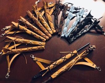 May include: A collection of leather wrapped wands with various colors and styles. The wands are tied with leather straps and have a decorative knot at the end. Some of the wands have a skull-shaped handle.