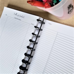 May include: A white notebook with black lines and a black spiral binding. The notebook has the words "Table of Contents" and "Topic & Description" printed on the cover. The notebook is open to a page with the words "Page #" and "Topic" printed at the top.