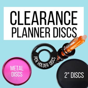 May include: A collection of planner discs including a bright pink metal disc, a black pen holder disc with white text, and a black 2-inch disc. The image features the text CLEARANCE PLANNER DISCS at the top, with an orange pen inserted into the discs.