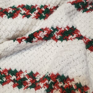 May include: A white crocheted blanket with red and green stripes. The blanket has a textured, plush appearance, with the stripes running diagonally across the surface. The red and green colors are interspersed with white, creating a festive, holiday-themed design.