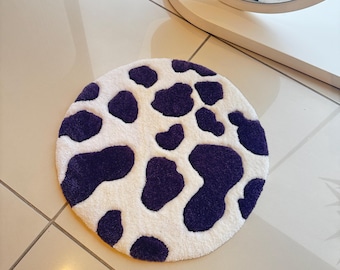 May include: A round, white rug with dark purple cow spots. The rug is on a tiled floor, and a mirror reflects the rug's design. The rug's design is a playful and whimsical addition to any room.