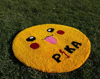 Può includere: Un tappeto rotondo giallo con un viso di cartone animato e la parola "PIKA" ricamata su di esso. Il viso ha occhi neri, una lingua rosa che sporge e guance rosse.