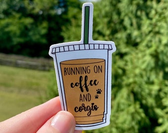 May include: A white sticker with a brown coffee cup design. The text on the sticker reads "Running on coffee and corgis" with paw prints.