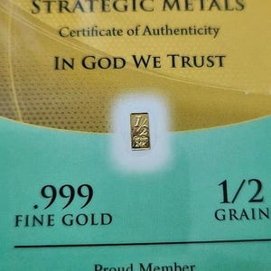 May include: A certificate of authenticity for a 1/2 grain .999 fine gold bar. The certificate is gold and green with the text "Benchmark Strategic Metals" and "In God We Trust". The certificate also includes the logo for the Jewelers Vigilance Committee.