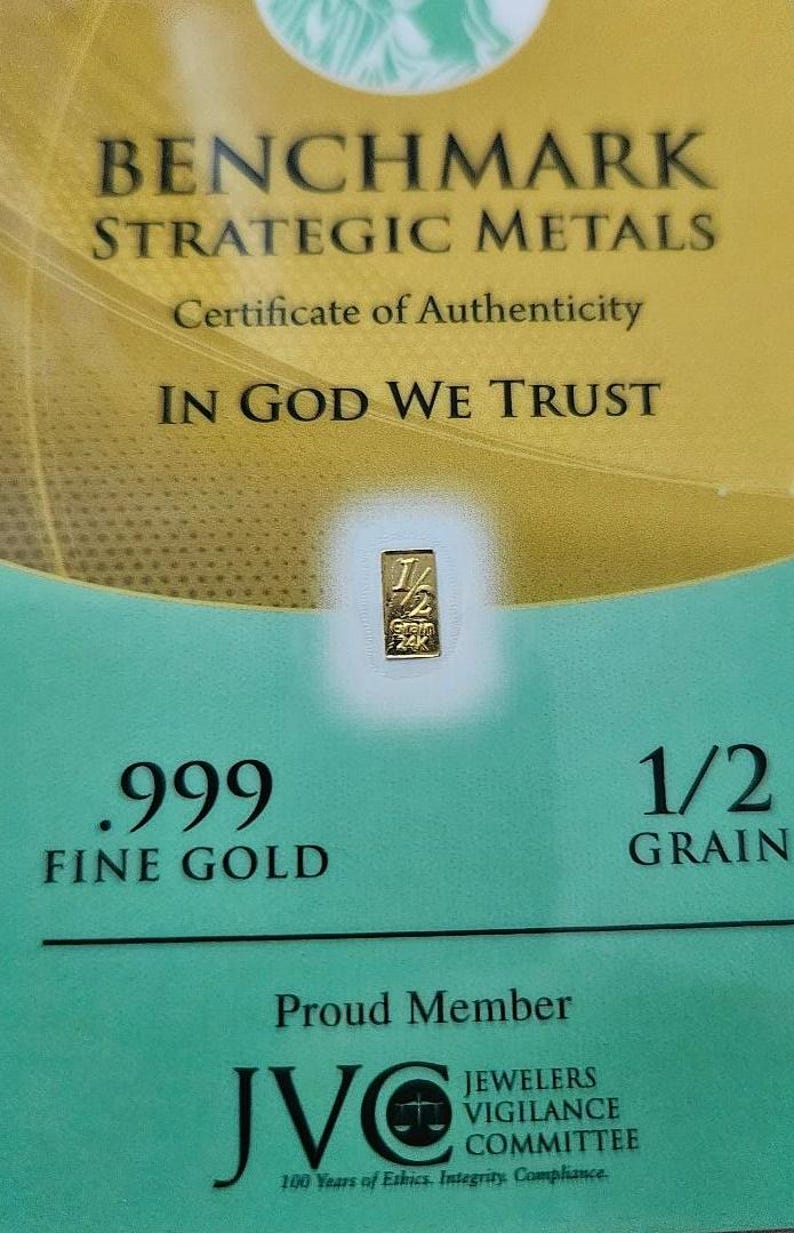 May include: A certificate of authenticity for a 1/2 grain .999 fine gold bar. The certificate is gold and green with the text "Benchmark Strategic Metals" and "In God We Trust". The certificate also includes the logo for the Jewelers Vigilance Committee.