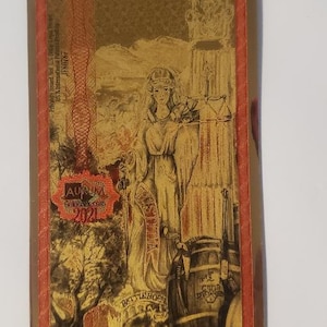 May include: A gold-colored paper bill with the text "Ten Nevada Goldbacks" and "Voluntary Local Currency". The bill features a woman in a white robe holding a scale, a barrel, and a tree. The text "1/100 Troy Ounce 24K Gold" and "For Circulation in Nevada" is printed at the bottom of the bill.