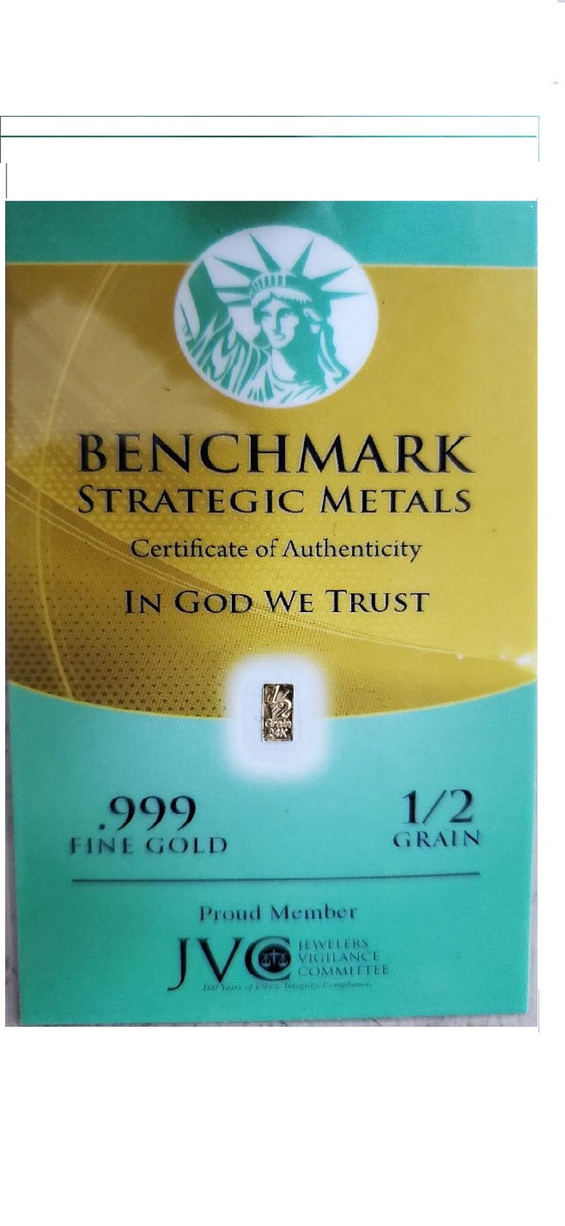 May include: A certificate of authenticity for a 1/2 grain .999 fine gold bar. The certificate features a green and gold design with the Statue of Liberty and the text "BENCHMARK STRATEGIC METALS" and "IN GOD WE TRUST". The certificate also includes the logo of the Jewelers Vigilance Committee (JVC).