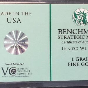 Puede incluir: Un certificado de autenticidad verde para una barra de oro fino de 1 grano de Benchmark Strategic Metals. El certificado presenta un sello hologr&aacute;fico con el n&uacute;mero 327850, una representaci&oacute;n de la Estatua de la Libertad y el texto "In God We Trust".