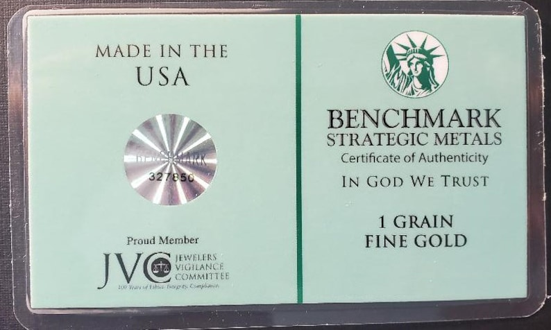 Puede incluir: Un certificado de autenticidad verde para una barra de oro fino de 1 grano de Benchmark Strategic Metals. El certificado presenta un sello hologr&aacute;fico con el n&uacute;mero 327850, una representaci&oacute;n de la Estatua de la Libertad y el texto "In God We Trust".