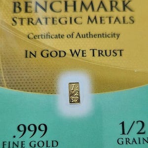 May include: A gold certificate of authenticity for a 1/2 grain .999 fine gold bar. The certificate features the text "BENCHMARK STRATEGIC METALS", "Certificate of Authenticity", "IN GOD WE TRUST", ".999 FINE GOLD", and "1/2 GRAIN".