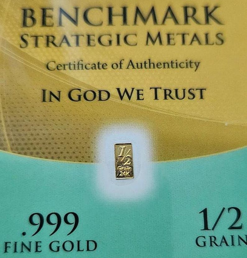 May include: A gold certificate of authenticity for a 1/2 grain .999 fine gold bar. The certificate features the text "BENCHMARK STRATEGIC METALS", "Certificate of Authenticity", "IN GOD WE TRUST", ".999 FINE GOLD", and "1/2 GRAIN".