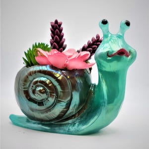 May include: A teal blue snail planter with a iridescent shell and a pink flower and green succulents inside.