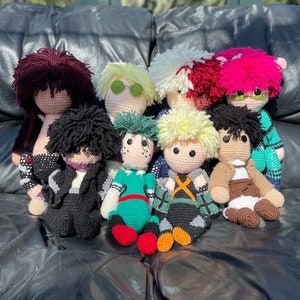 May include: A group of nine crocheted plush toys, each representing a different character. The toys are sitting on a black leather sofa. The characters are all wearing different outfits and have different hairstyles.