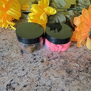 May include: Two small jars with black lids, one filled with a pink scrub and the other with a brown scrub. The jars are sitting on a brown speckled surface with yellow flowers in the background.