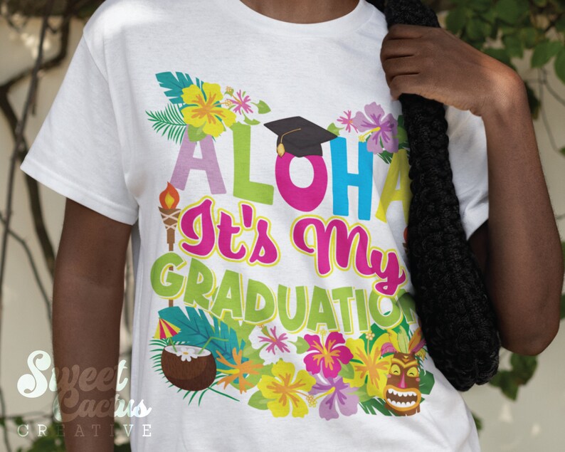 May include: White t-shirt with a colorful design featuring the text "Aloha It's My Graduation" with a graduation cap, tropical flowers, and a tiki mask.