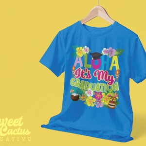 May include: A blue t-shirt with a colorful design that says "Aloha It's My Graduation" with a graduation cap, flowers, and a tiki mask.
