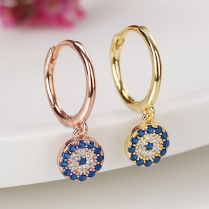 Pode incluir: Um par de brincos de argola dourados com um amuleto de olho turco azul e branco pendurado em cada argola.
