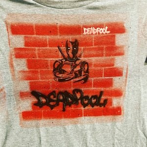 Pode incluir: Uma camiseta cinza com um design de parede de tijolo vermelho que apresenta uma ilustração em preto e branco do Deadpool. O texto "DEADPOOL" está escrito em branco na parede e em preto abaixo da ilustração.