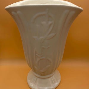 May include: Cream-colored ceramic vase with a flared top and a pedestal base. The vase features a floral relief design and vertical fluting. A decorative piece for home decor.
